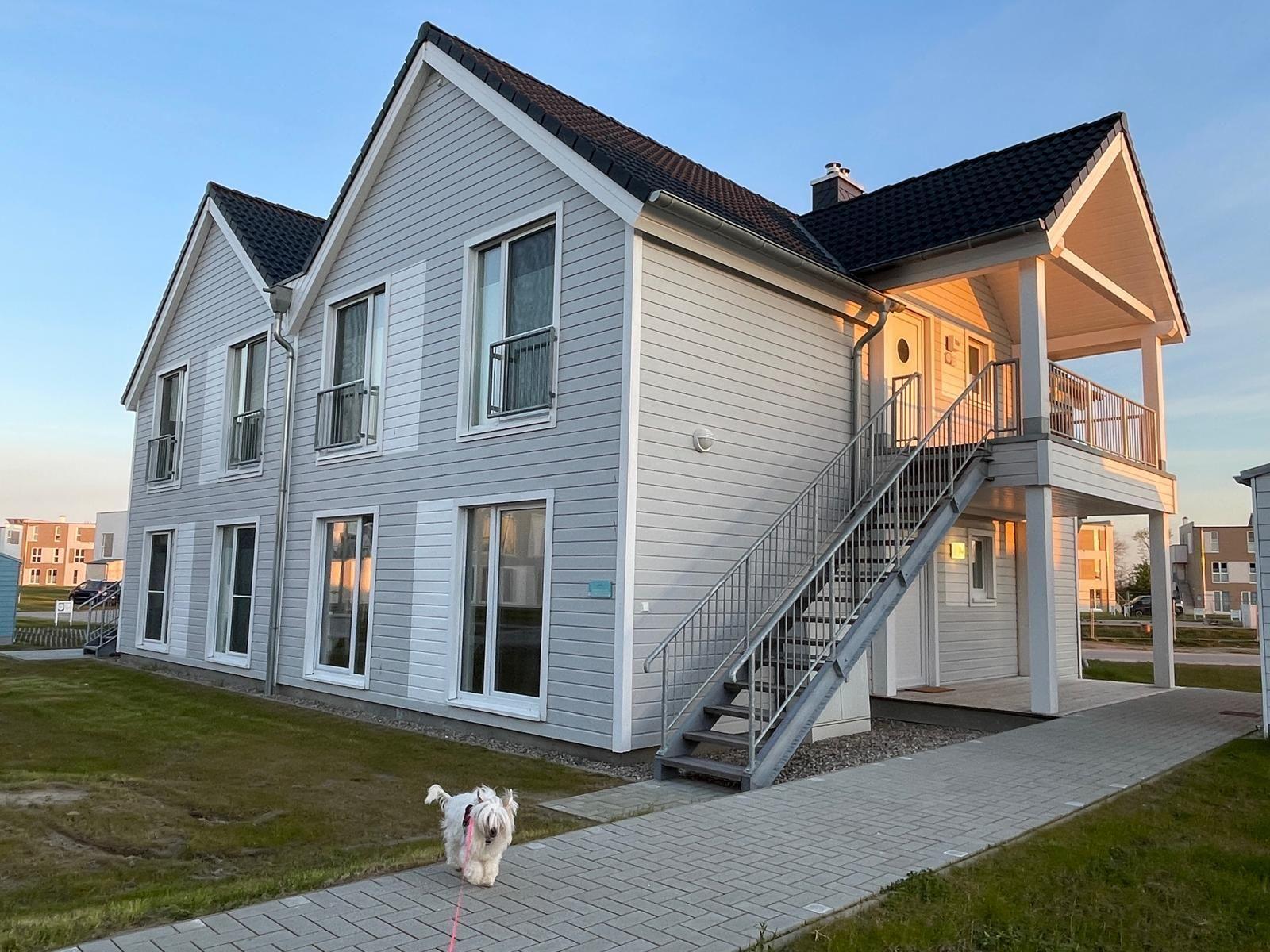 Zwei-Stöckiges Haus mit Balkon und Außentreppe. Ein weißer Hund läuft auf dem Weg.