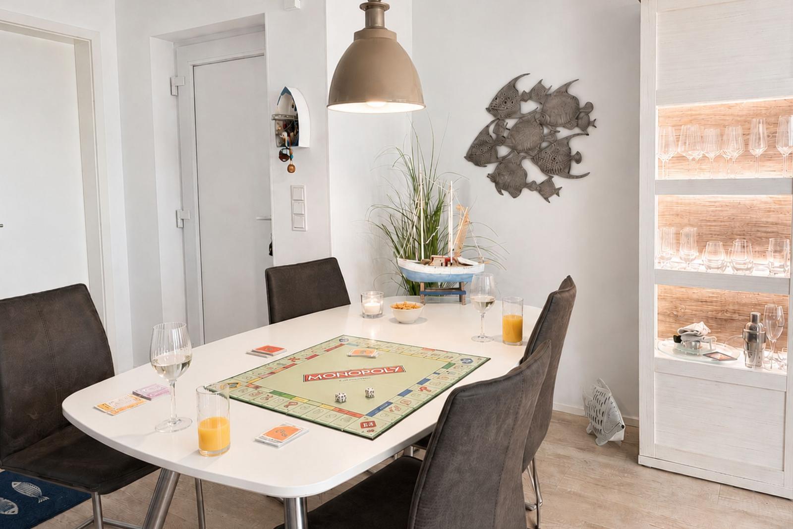 Esszimmer mit Tisch, Stühlen und Monopoly-Spiel. Wanddekoration und Getränkeflaschen sichtbar.