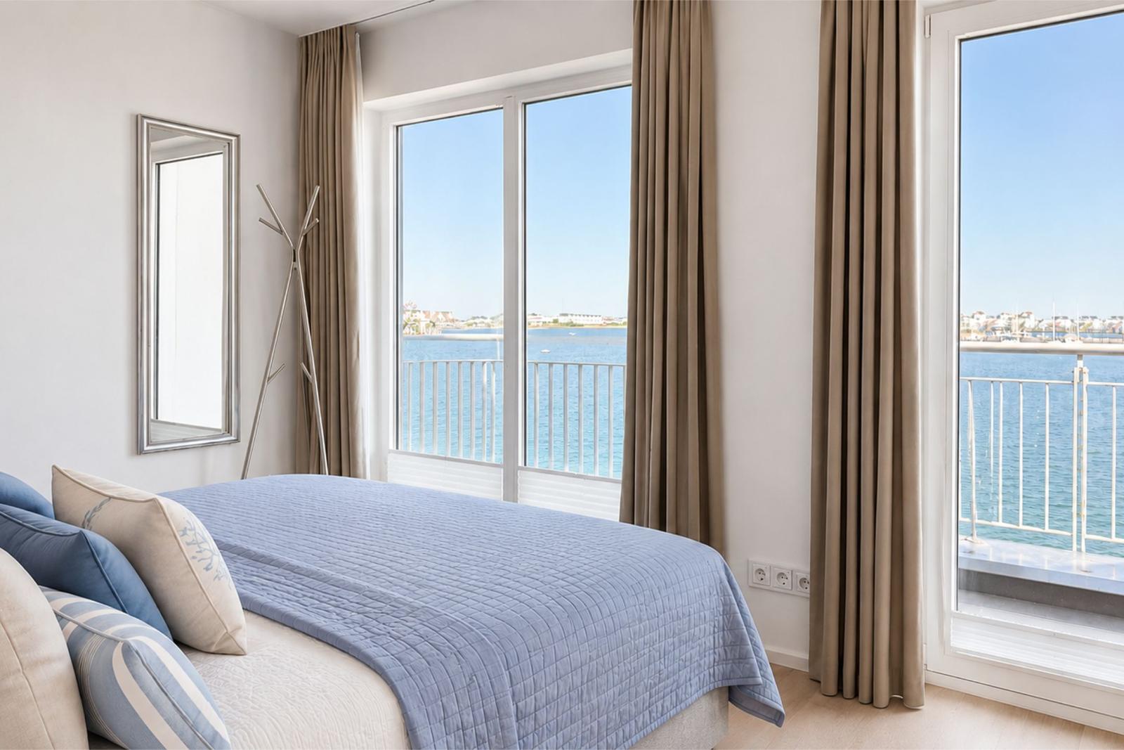 Schlafzimmer mit Blick aufs Wasser und Balkon. Bett, Spiegel und Gardinen sind sichtbar.