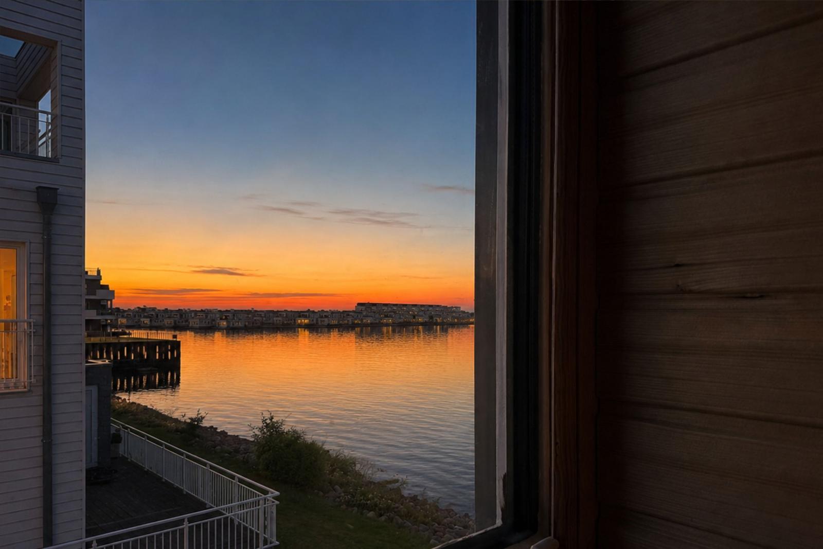 Aus dem Fenster blickt man auf einen See bei Sonnenuntergang mit gegenüberliegenden Häusern.