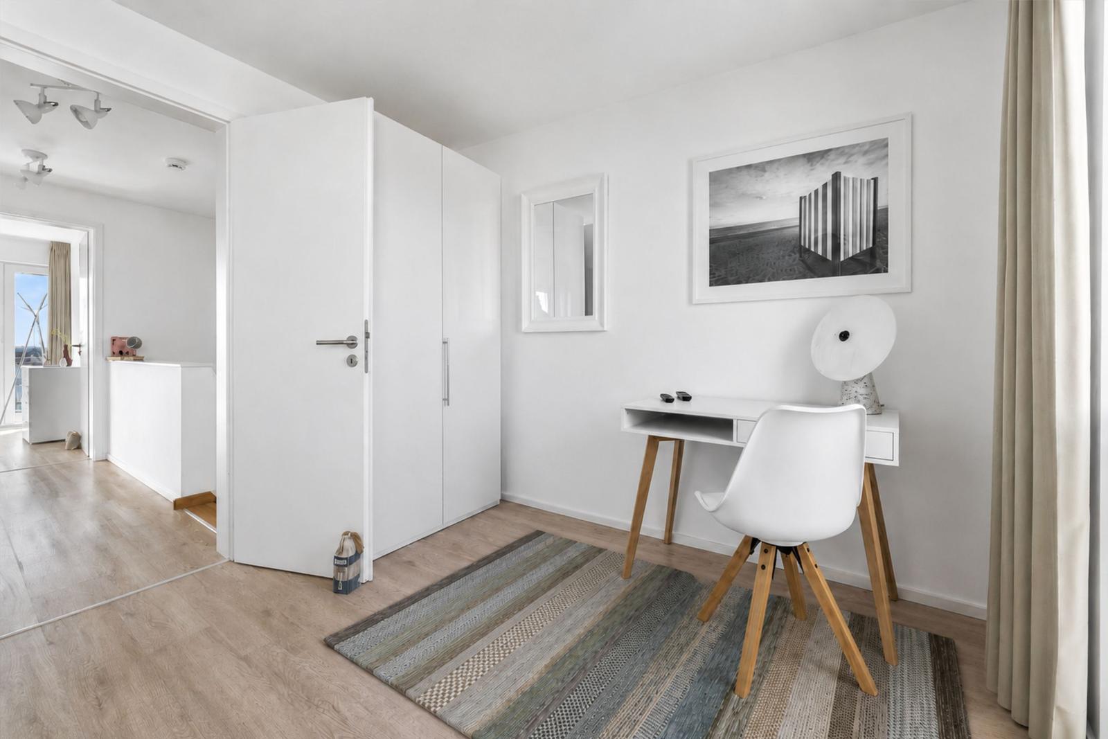 Ein Zimmer mit Schreibtisch, Stuhl und Kleiderschrank. Wandbild und Teppich sind sichtbar.