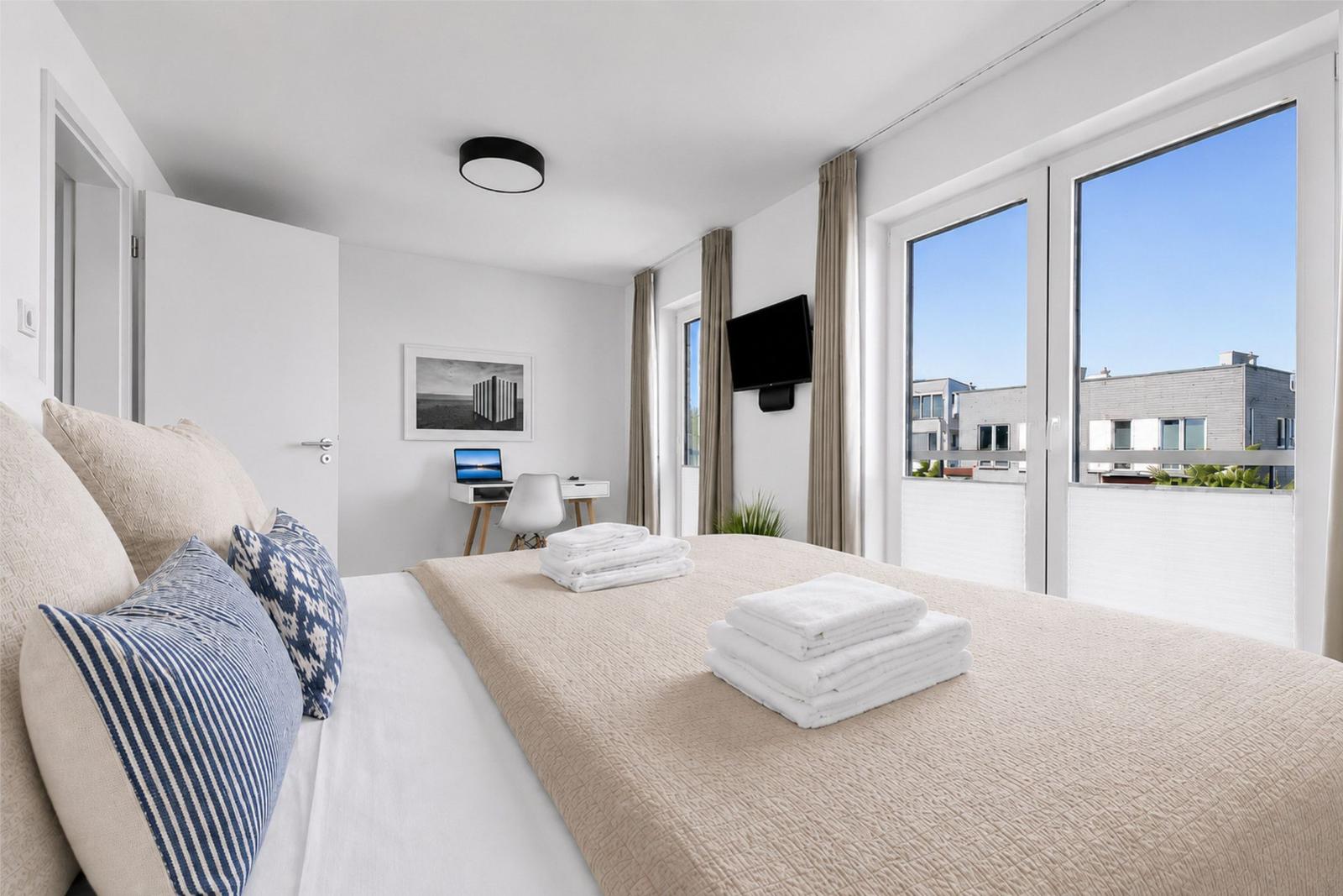 Schlafzimmer mit Bett, Schreibtisch und TV. Große Fenster mit Blick auf Balkon.
