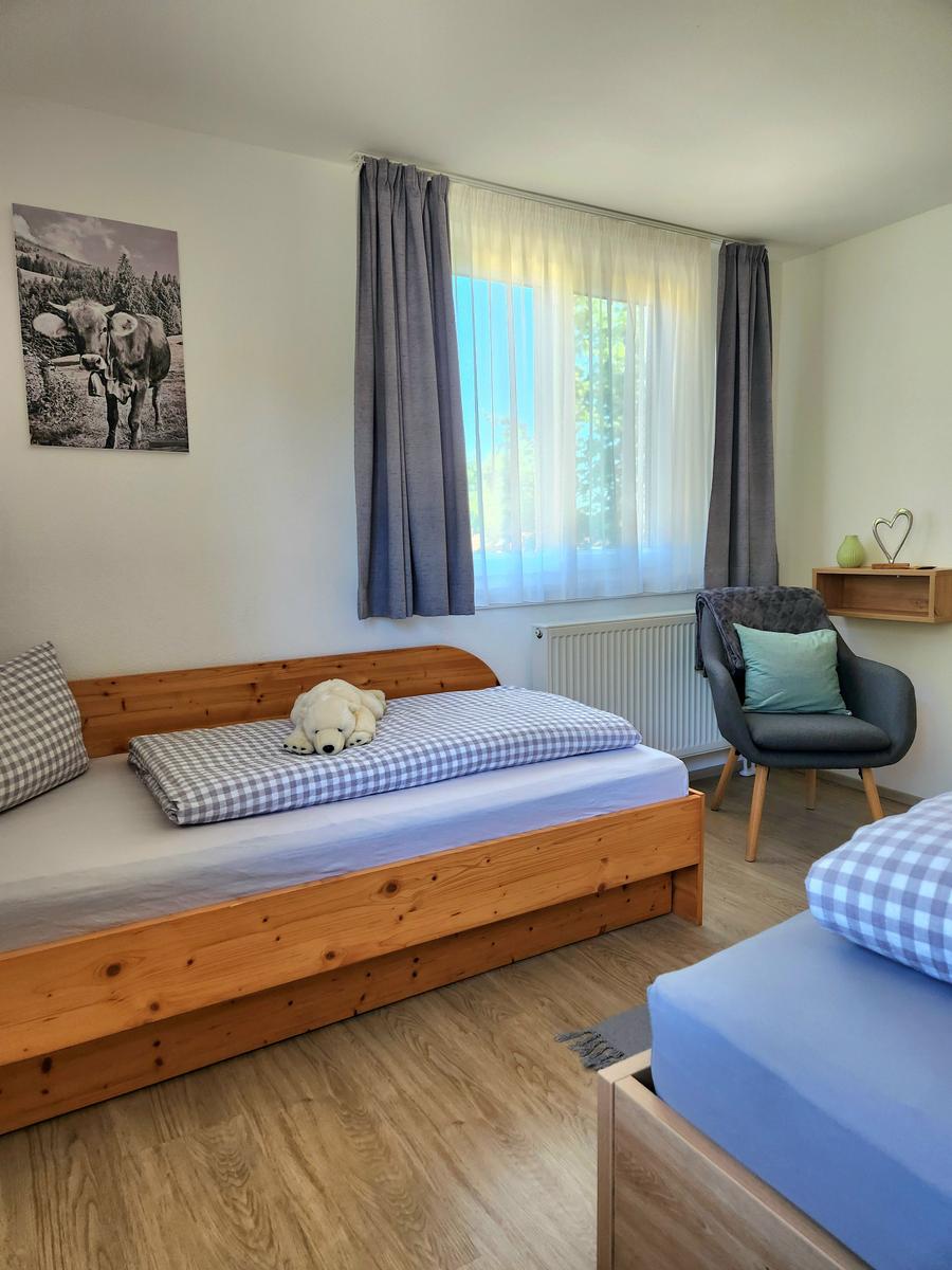 Zimmer mit zwei Betten, Fenster mit Vorhängen und Stuhl mit Kissen.