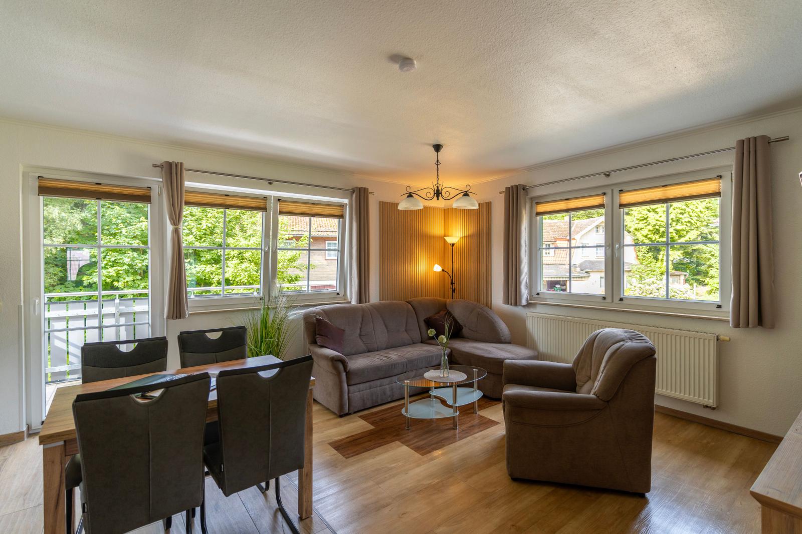 Wohn- und Essbereich mit Sofa, Sessel und Tisch. Fenster mit Blick auf Grün.