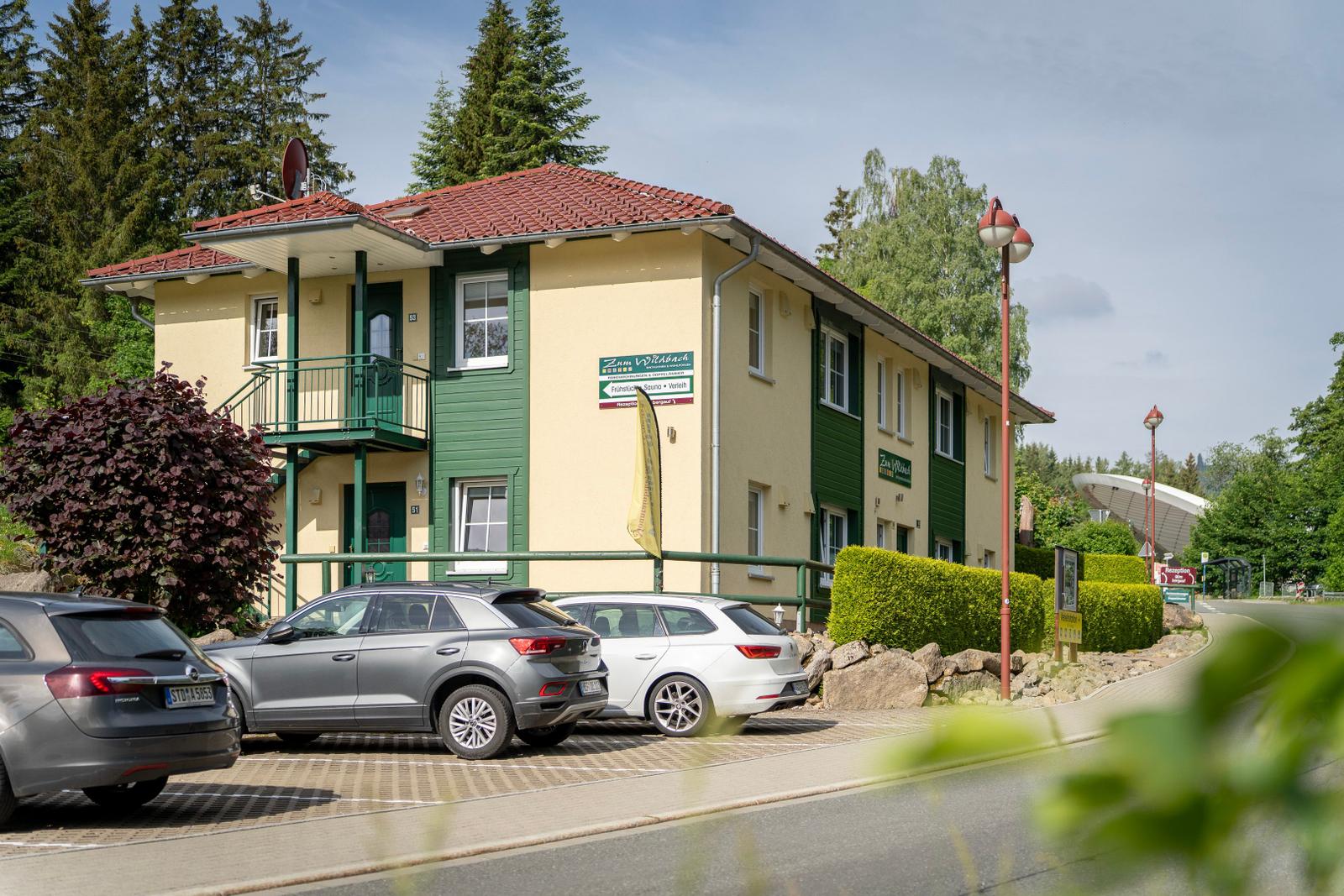 Zwei-Stockwerksgebäude mit Terrasse, Parkplatz und grüner Umgebung.