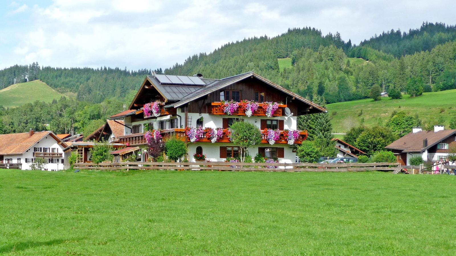 Haus mit Blumenbalkonen, grünen Hügeln und Wald im Hintergrund.