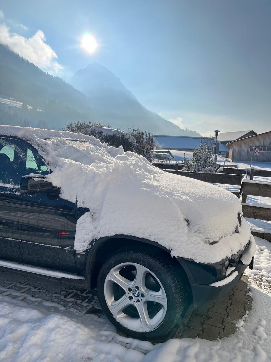 Schneebedecktes Auto vor verschneiten Bergen und einem Holzhaus.