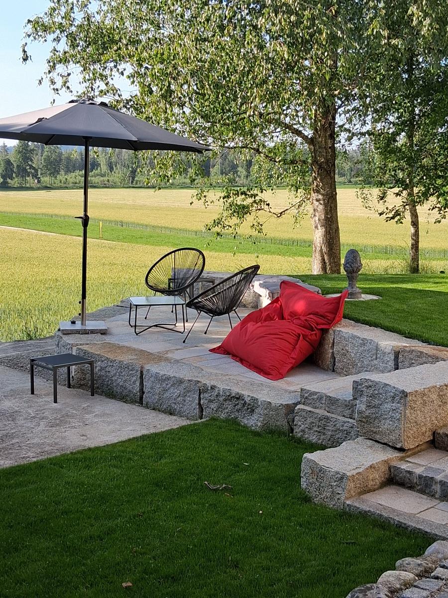 Außenbereich mit Sonnenschirm, zwei Stühlen und rotem Sitzkissen auf Steinplattform unter Baum.