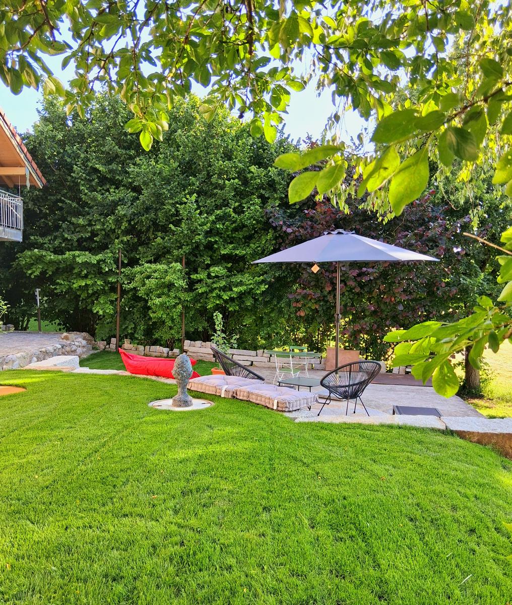 Grüner Garten mit Liegefläche, Sonnenschirm und Skulptur unter Bäumen.