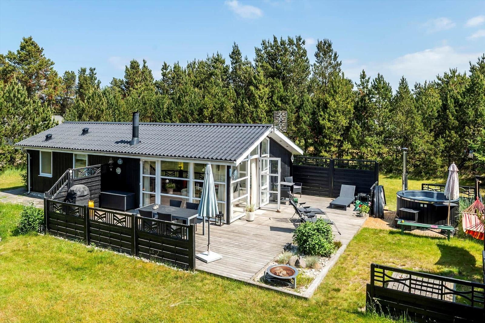 Ein dunkel verputztes Ferienhaus mit weißer Tür und Terrasse. Umgeben von Gras und Bäumen unter blauem Himmel.