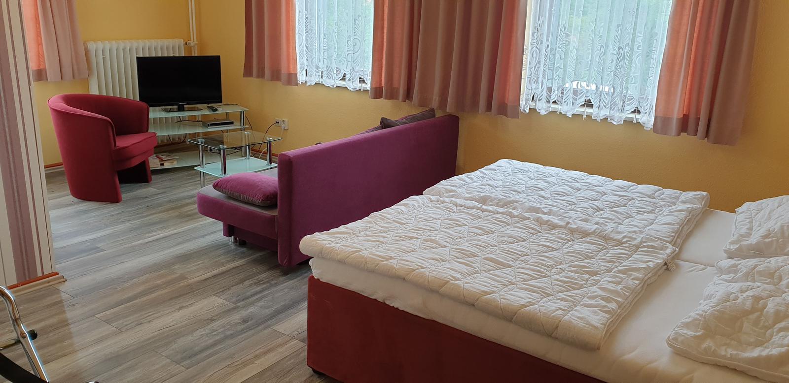 Zimmer mit Bett, Sofa, Sessel und Fernseher. Fenster mit Vorhängen.