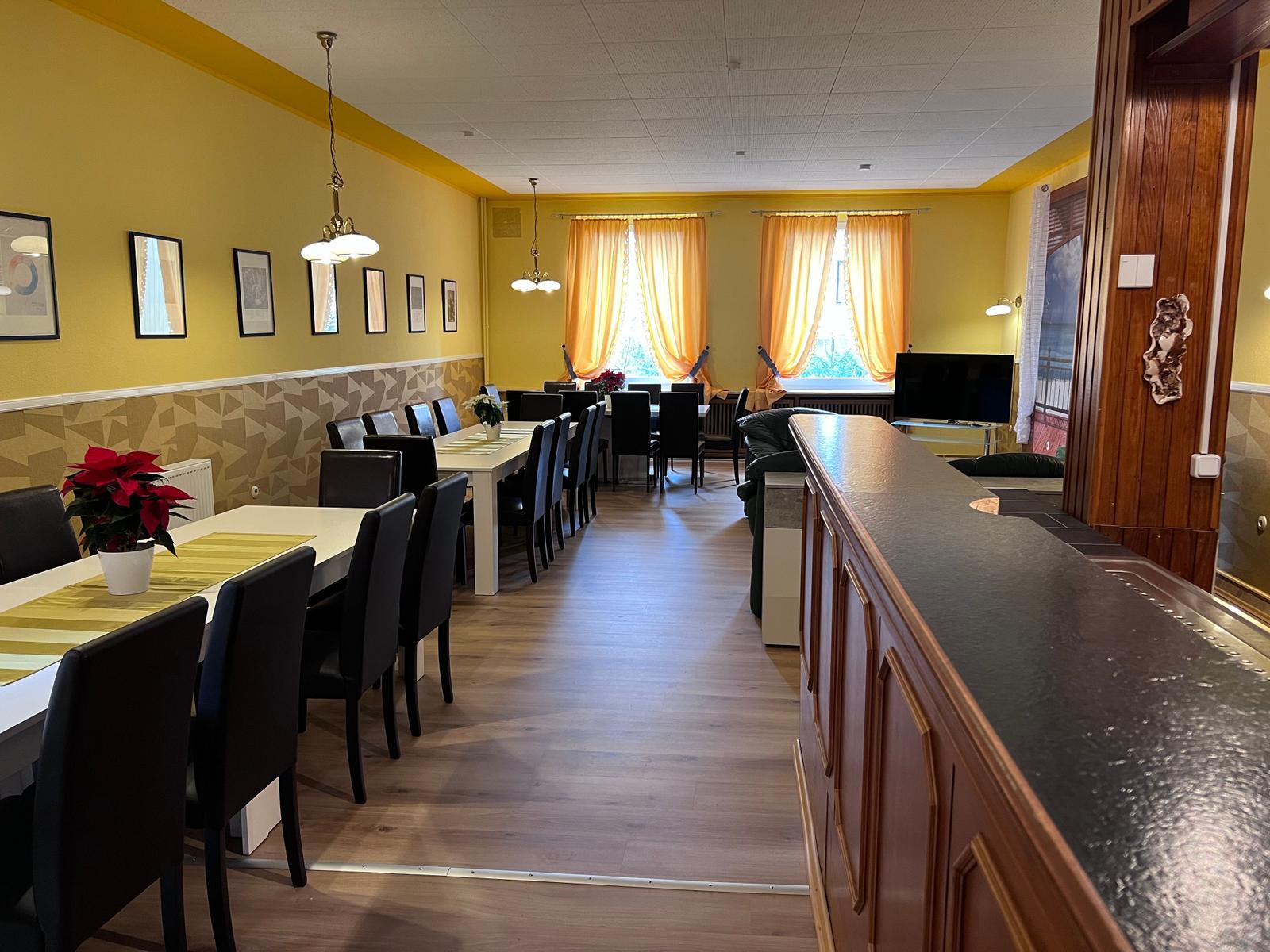 Großer Raum mit Essbereich, TV und Bar. Gelbe Wände, Holzboden und Fenster mit Vorhängen.