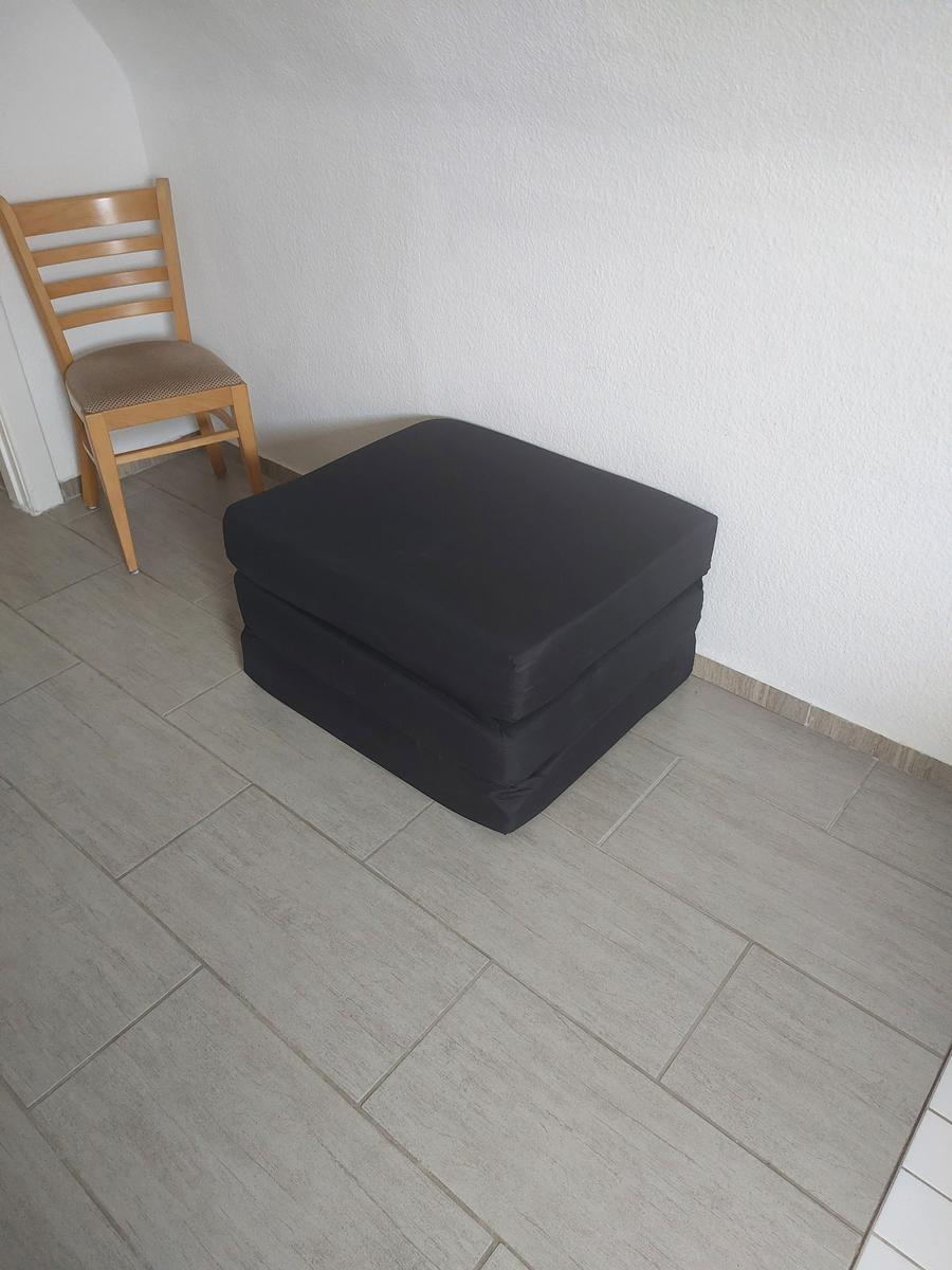 Black ottoman and wooden chair in a room with light walls and tiled floor.