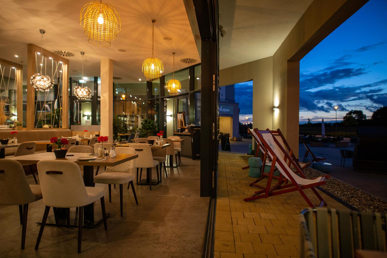 Innenraum mit Tischen und Stühlen, draußen Liegestühle und Terrasse bei Dämmerung.