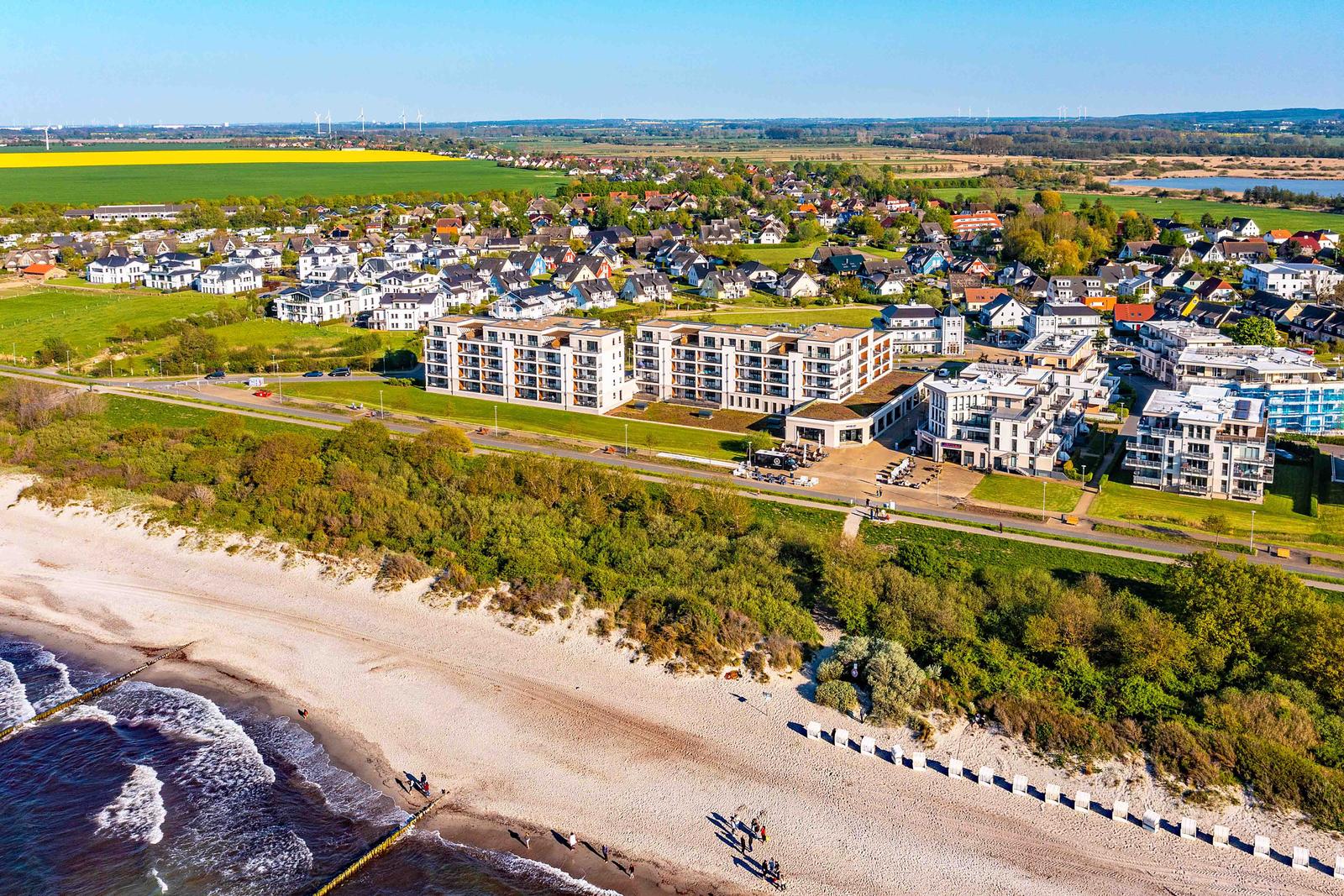 Strand mit Dünen, modernen Gebäuden und grünen Feldern im Hintergrund.