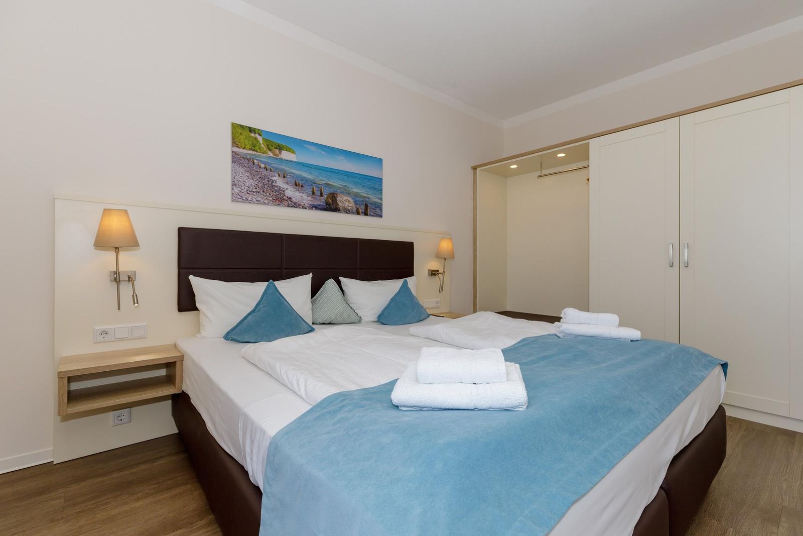 Schlafzimmer mit Doppelbett, Nachttischen und Kleiderschrank. Wandbild zeigt Küstenlandschaft.