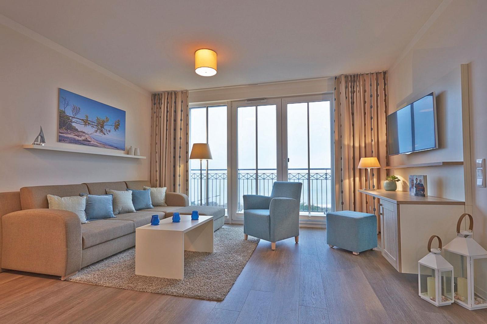 Wohnzimmer mit Sofa, Sessel und TV. Balkon mit Aussicht. Holzboden und Teppich.