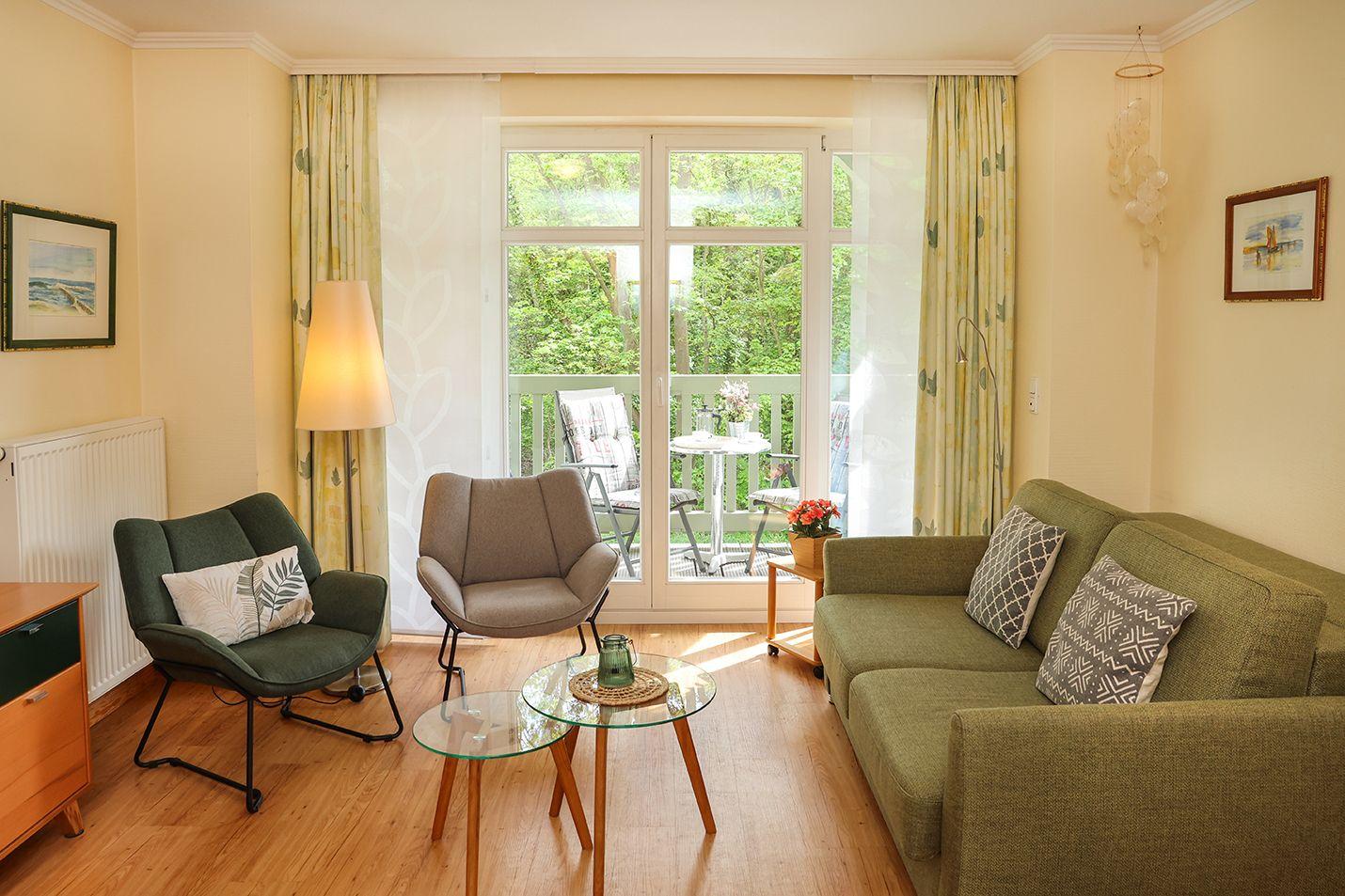 Wohnzimmer mit Sofa, zwei Sesseln und Glas-Tisch. Durch Fenster Blick auf Balkon mit Garten.