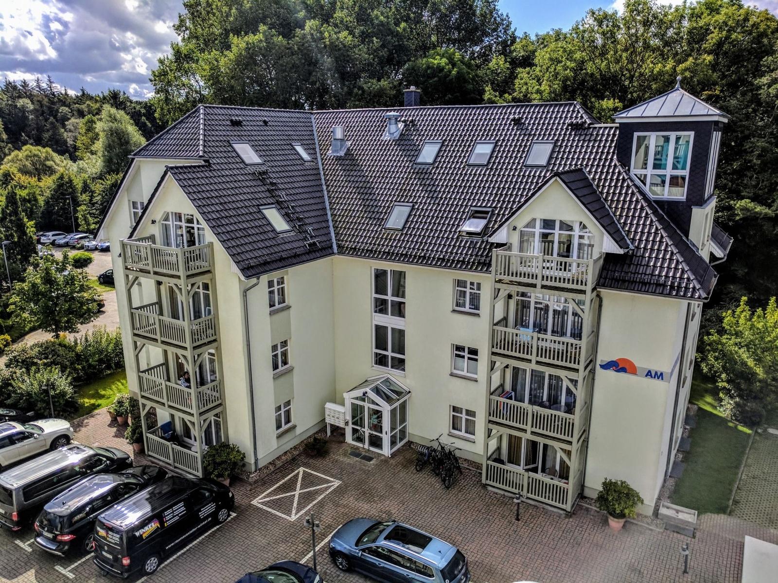 Ein mehrstöckiges Gebäude mit Balkonen und Dachfenstern, umgeben von Bäumen und einem Parkplatz.