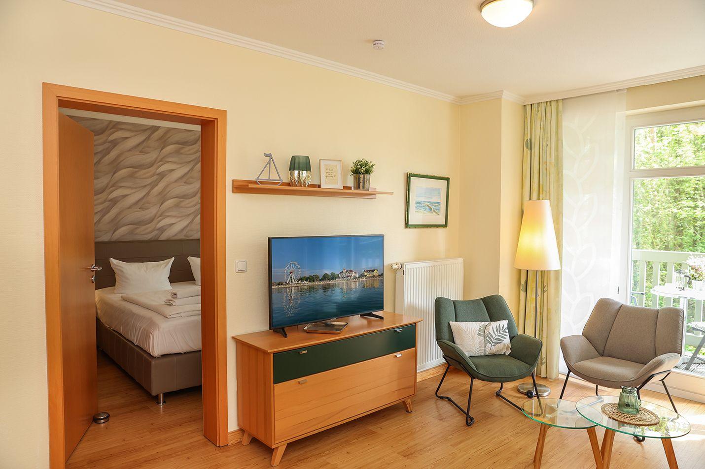 Wohnbereich mit TV, zwei Sesseln und Blick auf Balkon. Schlafzimmer mit Bett ist sichtbar.