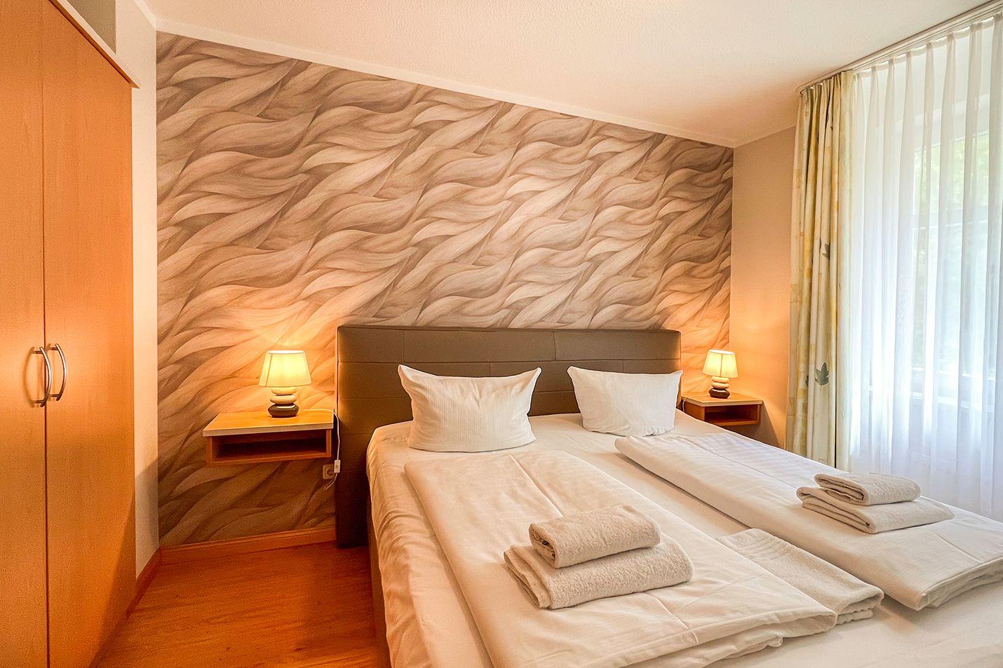 Schlafzimmer mit Bett, Nachttischen und Wandverkleidung. Holzfußboden und Fenster mit Vorhängen.