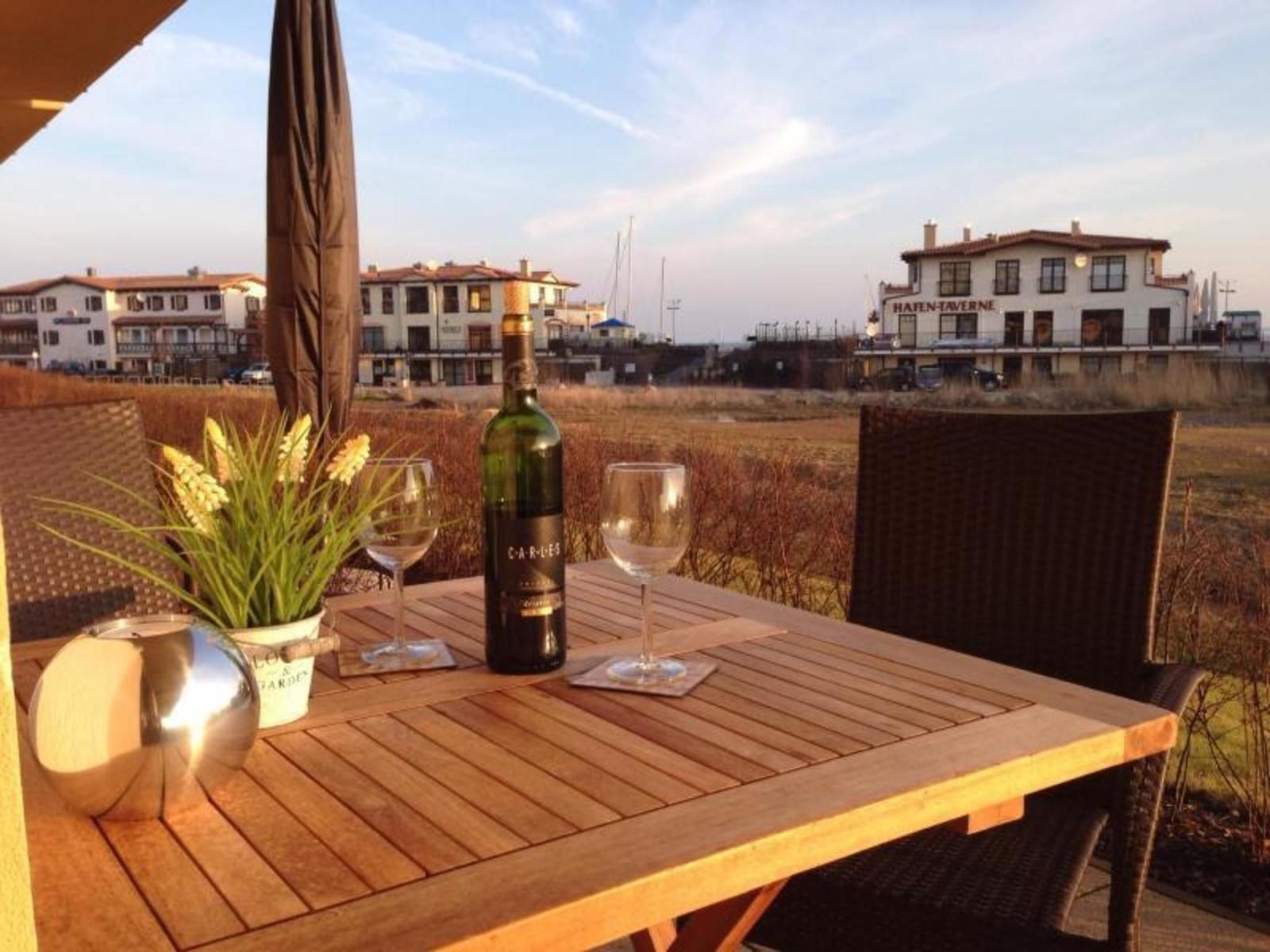 Holztisch mit Weinflasche und Gläsern auf Terrasse. Blick auf Hafen und Gebäude.