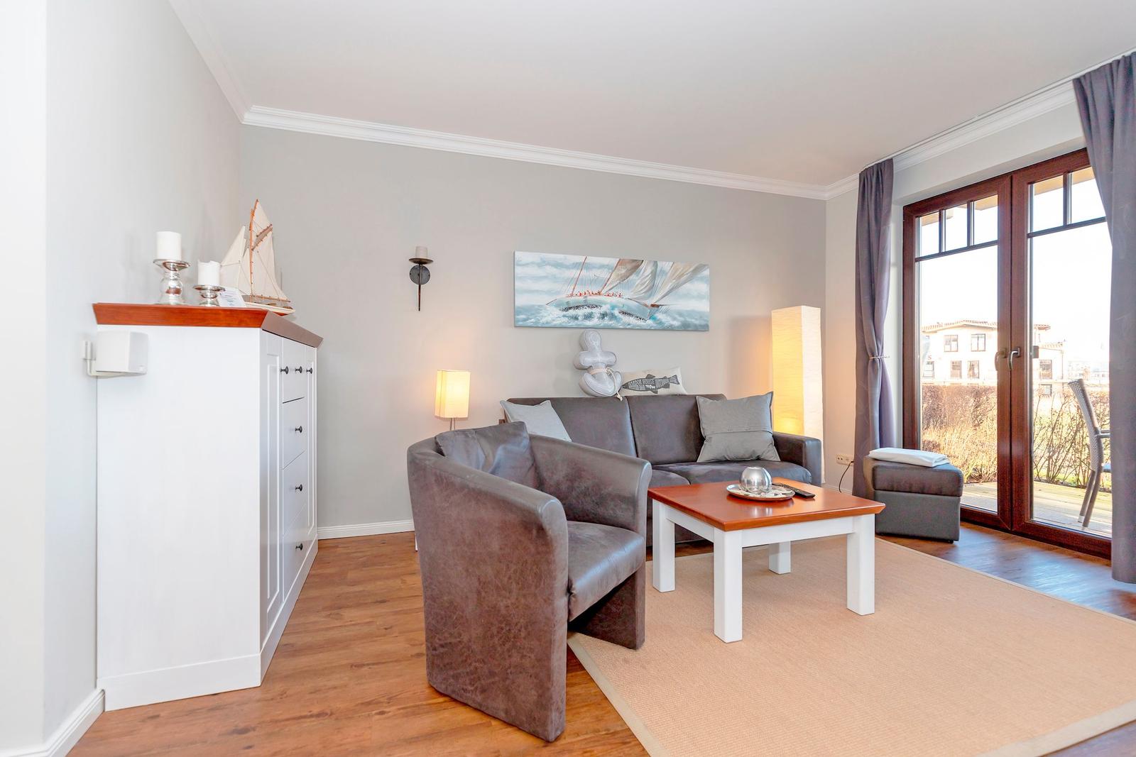 Wohnzimmer mit Sofa, Sessel und Tisch. An der Wand hängt ein Segelbootbild. Durch die Glastür sieht man draußen.
