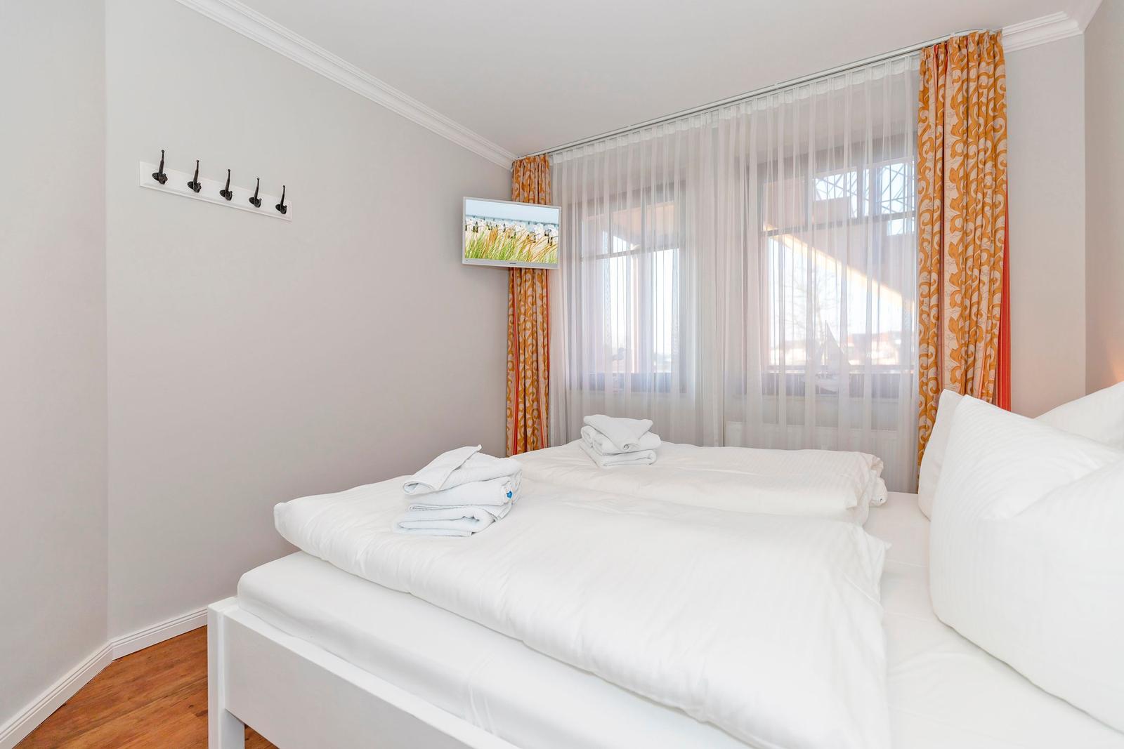 Schlafzimmer mit weißem Bett, orangefarbenen Vorhängen und Wandfernseher.