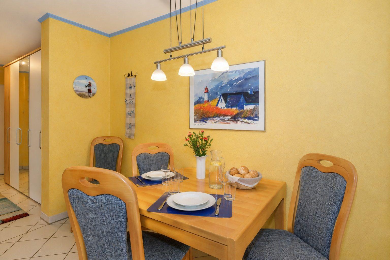 Esszimmer mit Holztisch, vier Stühlen und gelben Wänden. An der Wand hängt ein Bild mit Leuchten.