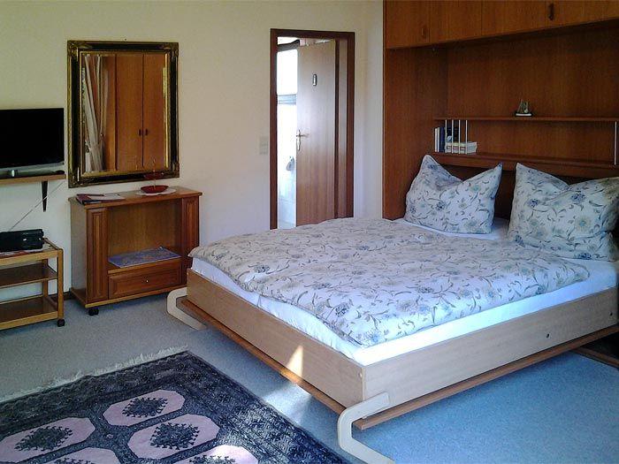 Schlafzimmer mit Bett, Schrank und Fernseher. Bodenbelag und Teppich sind sichtbar.