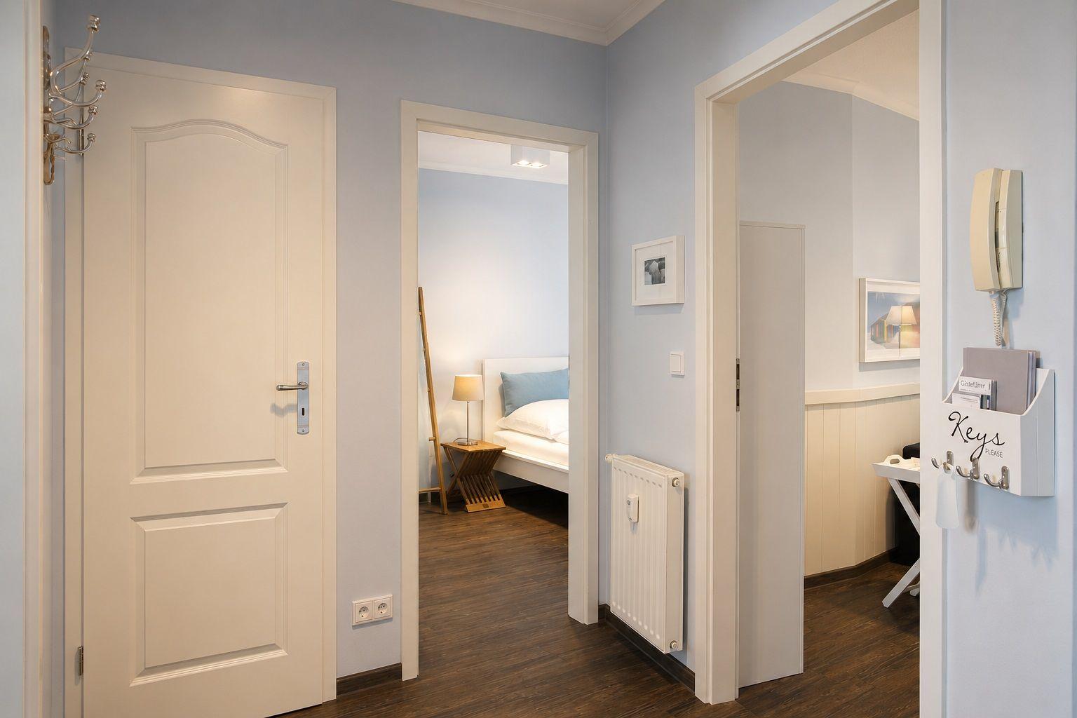 Flur mit Schlafzimmer und Arbeitsbereich. Tür, Heizung, Wandbild und Schlüsselhalter sichtbar.
