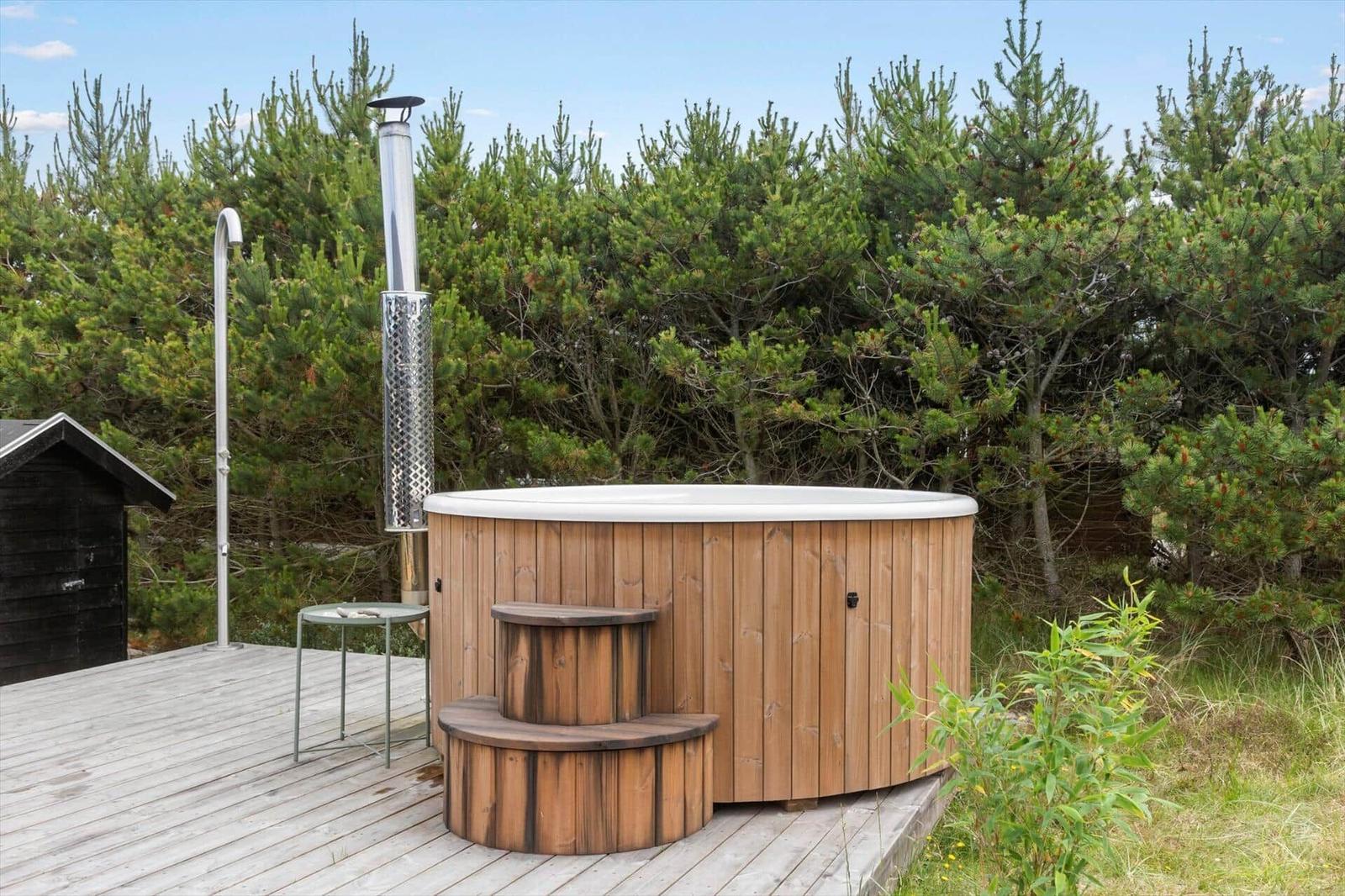 Backyard with wooden terrace, lounge chair, and hot tub between grass and trees.
