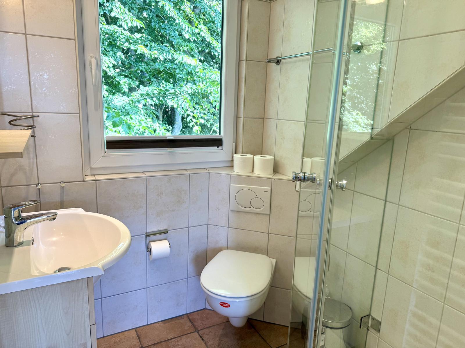 Badezimmer mit Waschbecken, WC unter Fenster und Glasdusche. Terrakotta-Boden.
