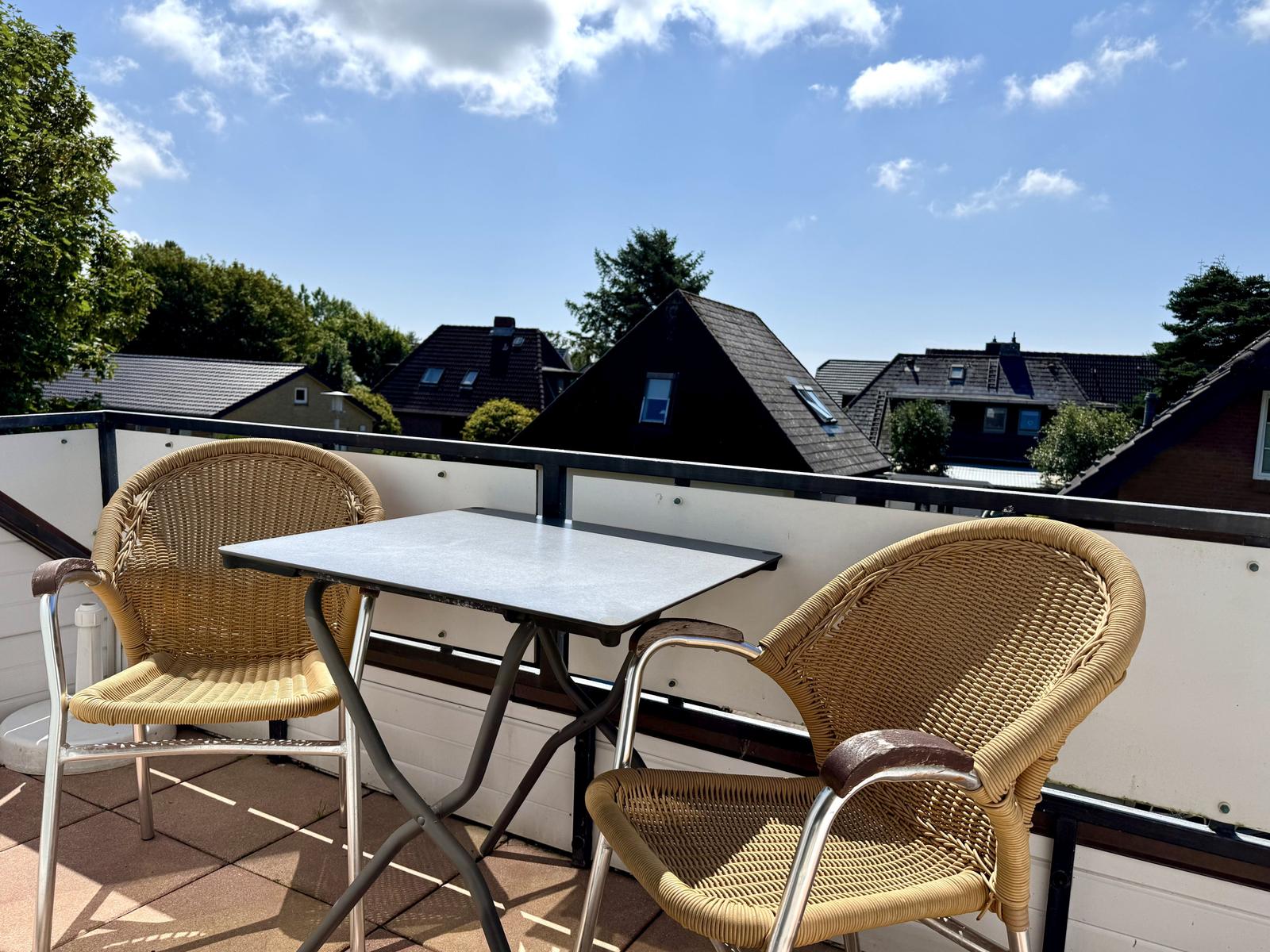 Balkon mit zwei Korbstühlen und Tisch. Blick auf Dächer und Bäume.