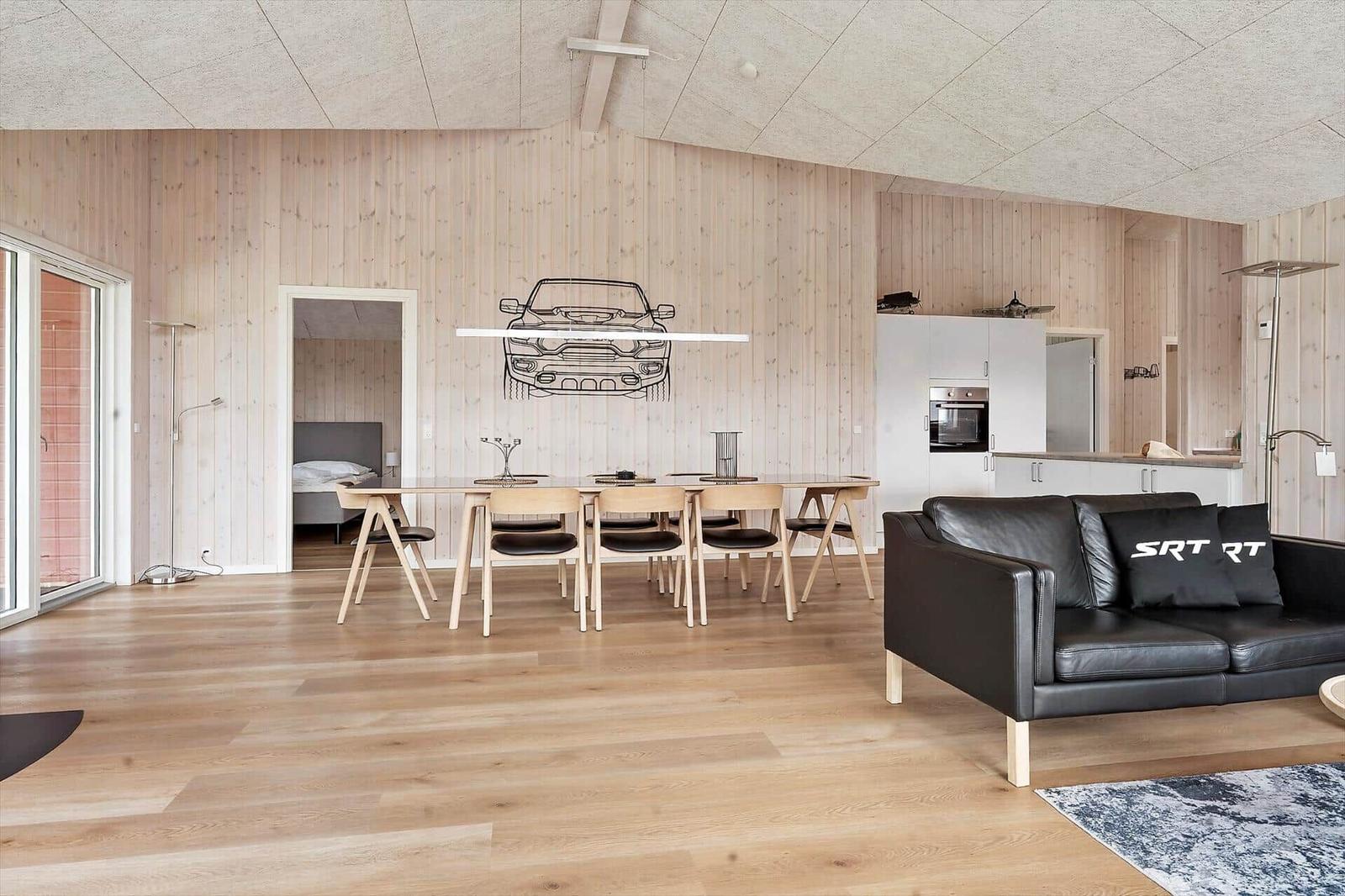 Living area with black leather furniture, wooden floor, and large window. Kitchen in the background.