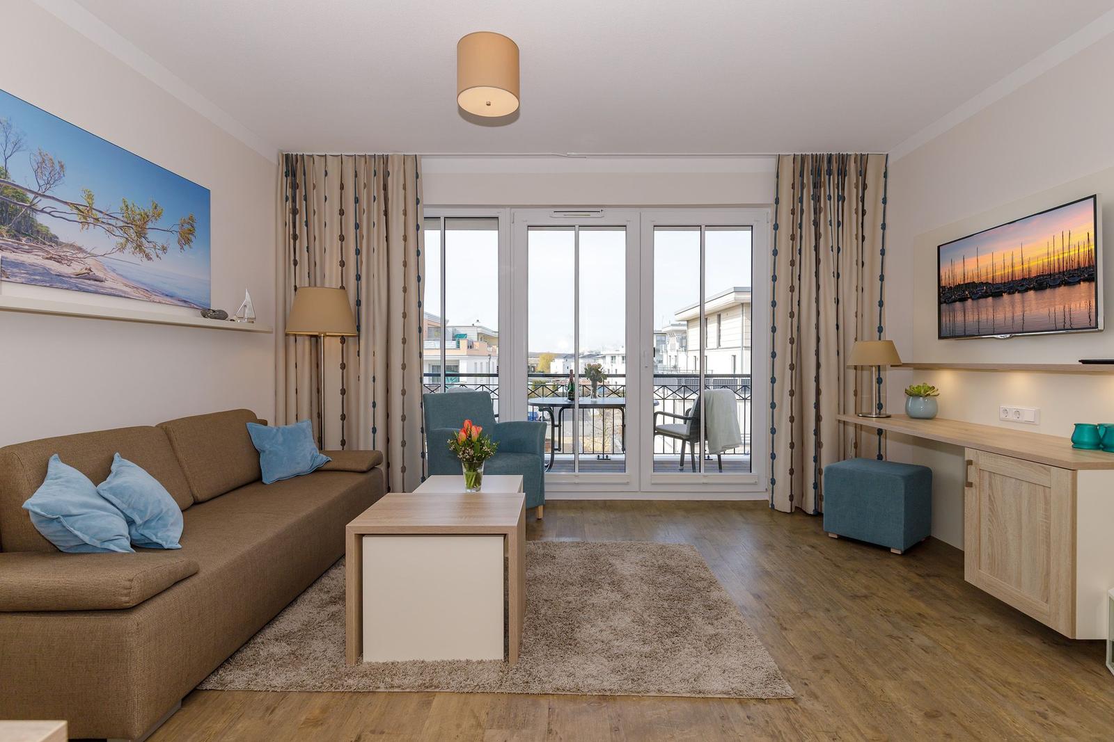 Wohnzimmer mit Sofa, Couchtisch und Balkon. Fernseher und Gardinen sichtbar.