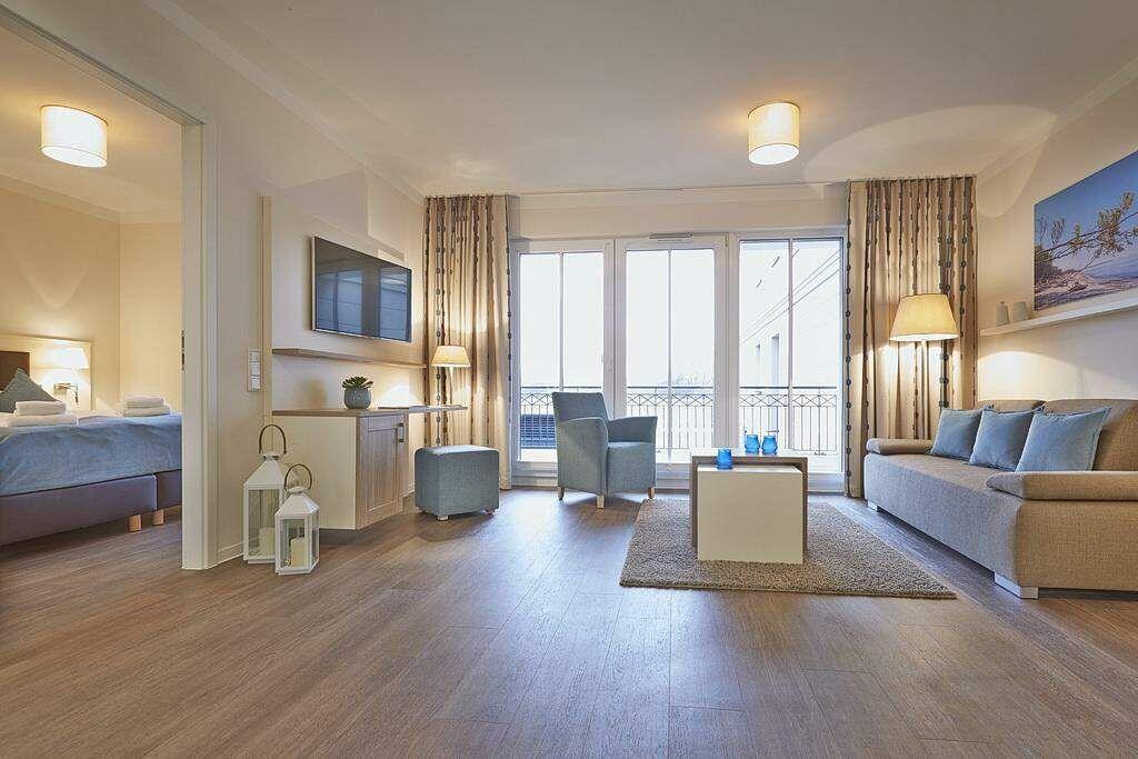 Wohnbereich mit Sofa, Sessel und Balkon. Schlafzimmer im Hintergrund.