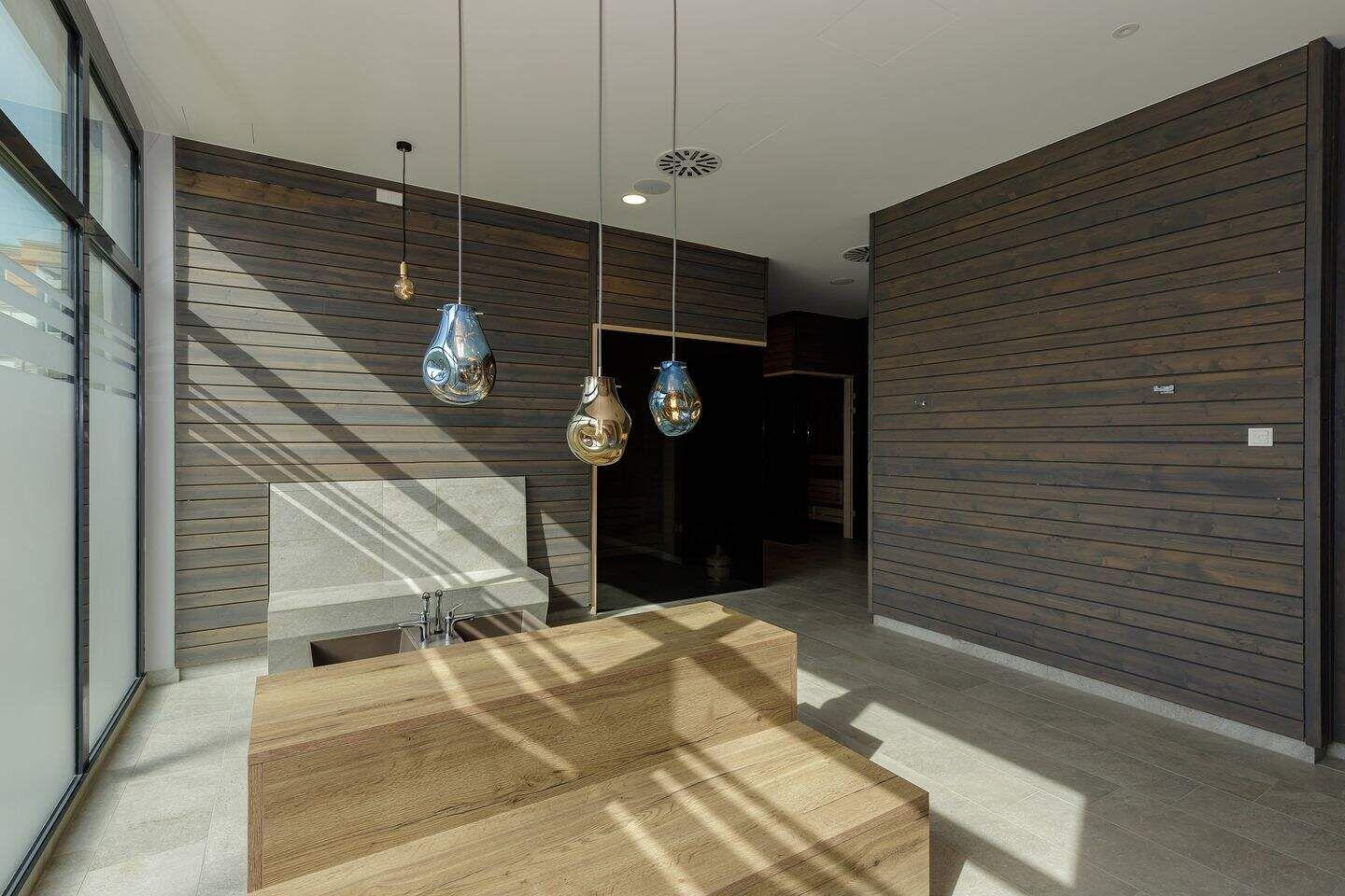 Raum mit Holzwand, Holzbank und hängenden Lampen. Große Fenster und Steinwaschbecken.