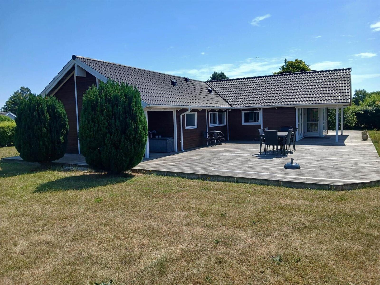 Moderne Villa mit großem Holzterrasse und Glasfront. Umgeben von Grün und Bäumen.