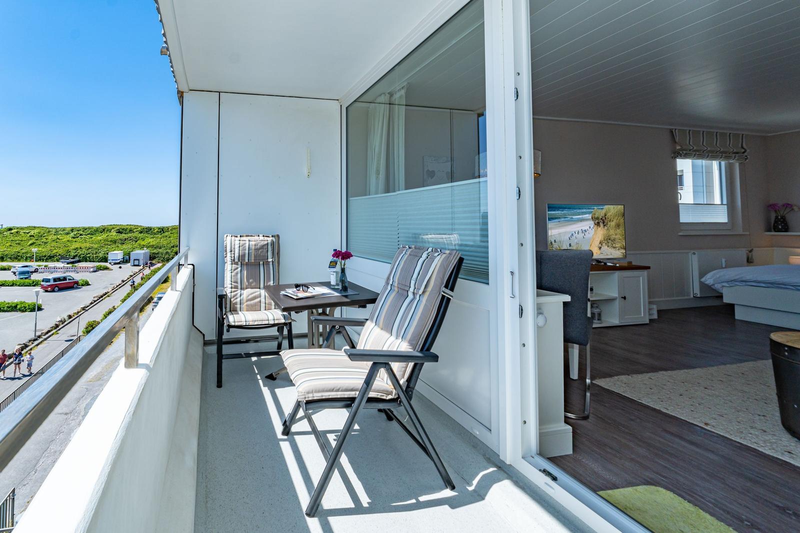 Balcony with two chairs and table, overlooking parking lot and greenery.