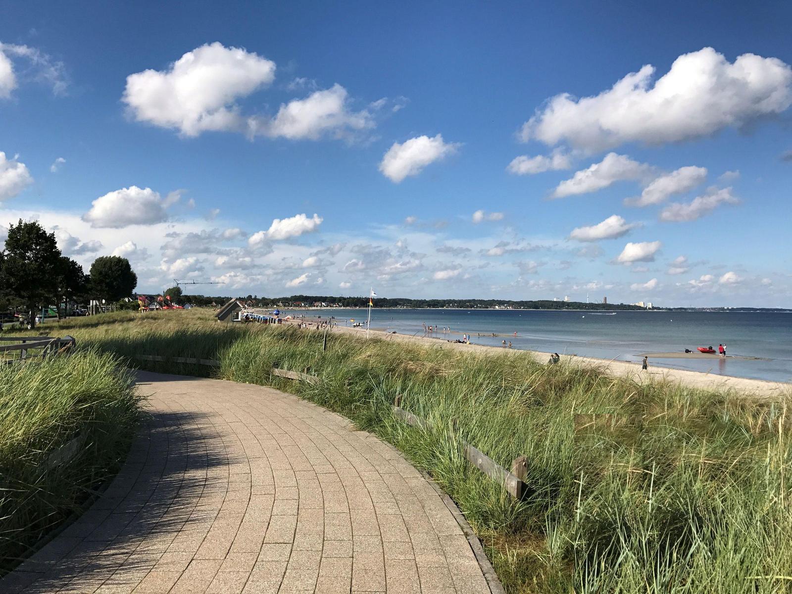 Grüne Dünen mit Blick auf Strand und Segelboot unter blauem Himmel.
