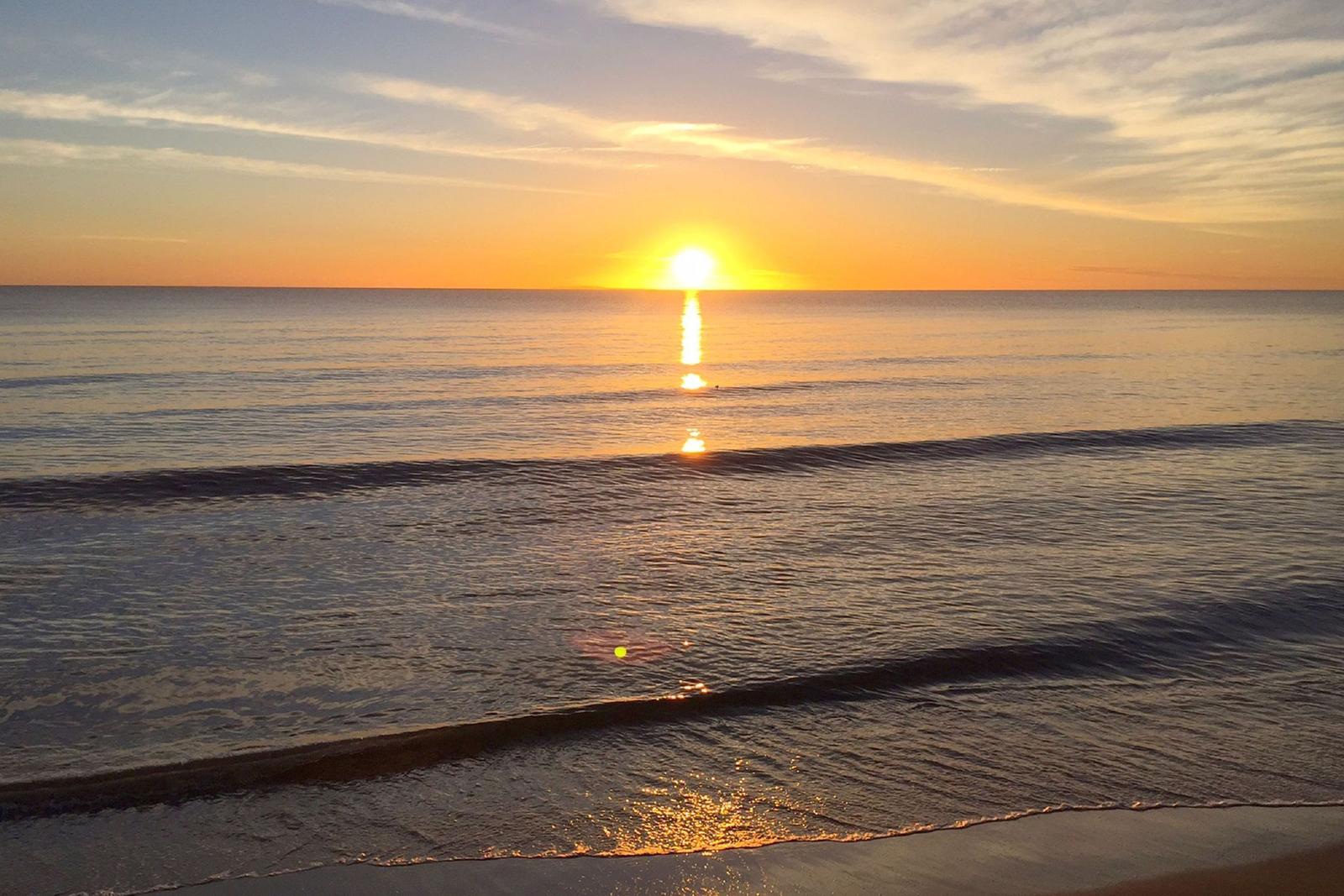 Strand mit sanften Wellen und Dünen am Horizont bei Sonnenuntergang.