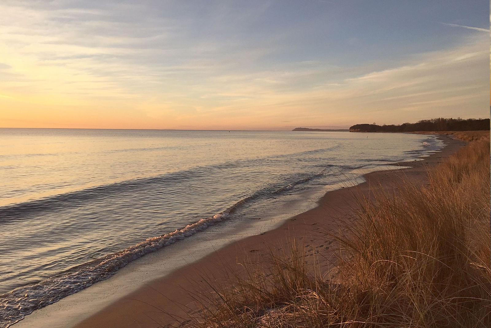 Strand mit Sand, Gras und Steinen am Wasser unter blauem Himmel mit Wolken.