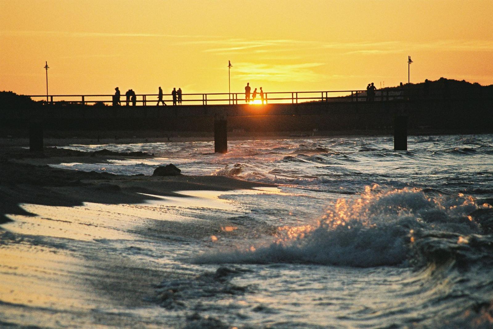 Holzsteg führt zum Strand mit Sonnenschirmen und Badegästen am Meer.
