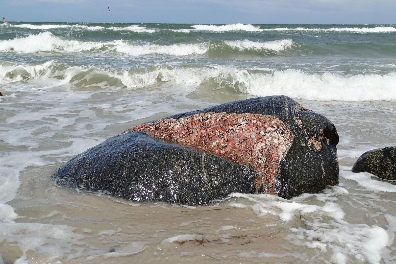 Strand mit Wellen, Felsen und einem Holzstamm im Vordergrund.