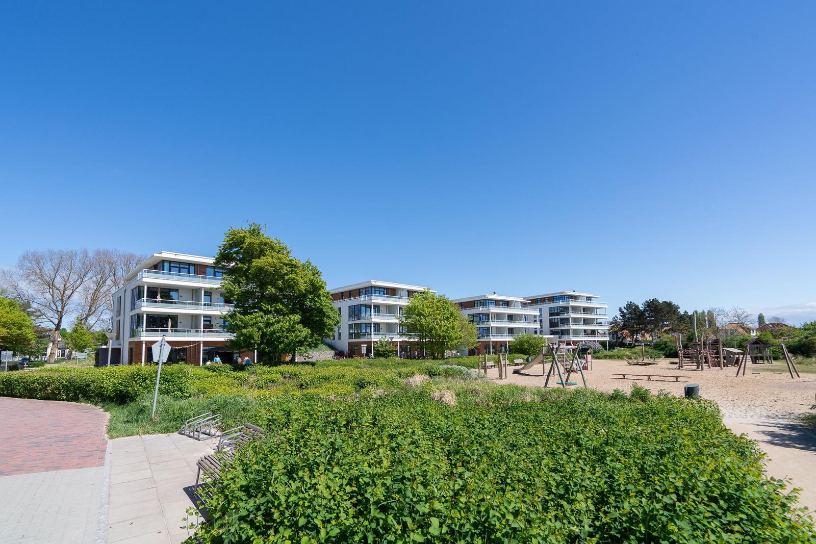 Moderne Wohngebäude mit Balkonen stehen unter blauem Himmel. Grasfläche und Bäume im Vordergrund.