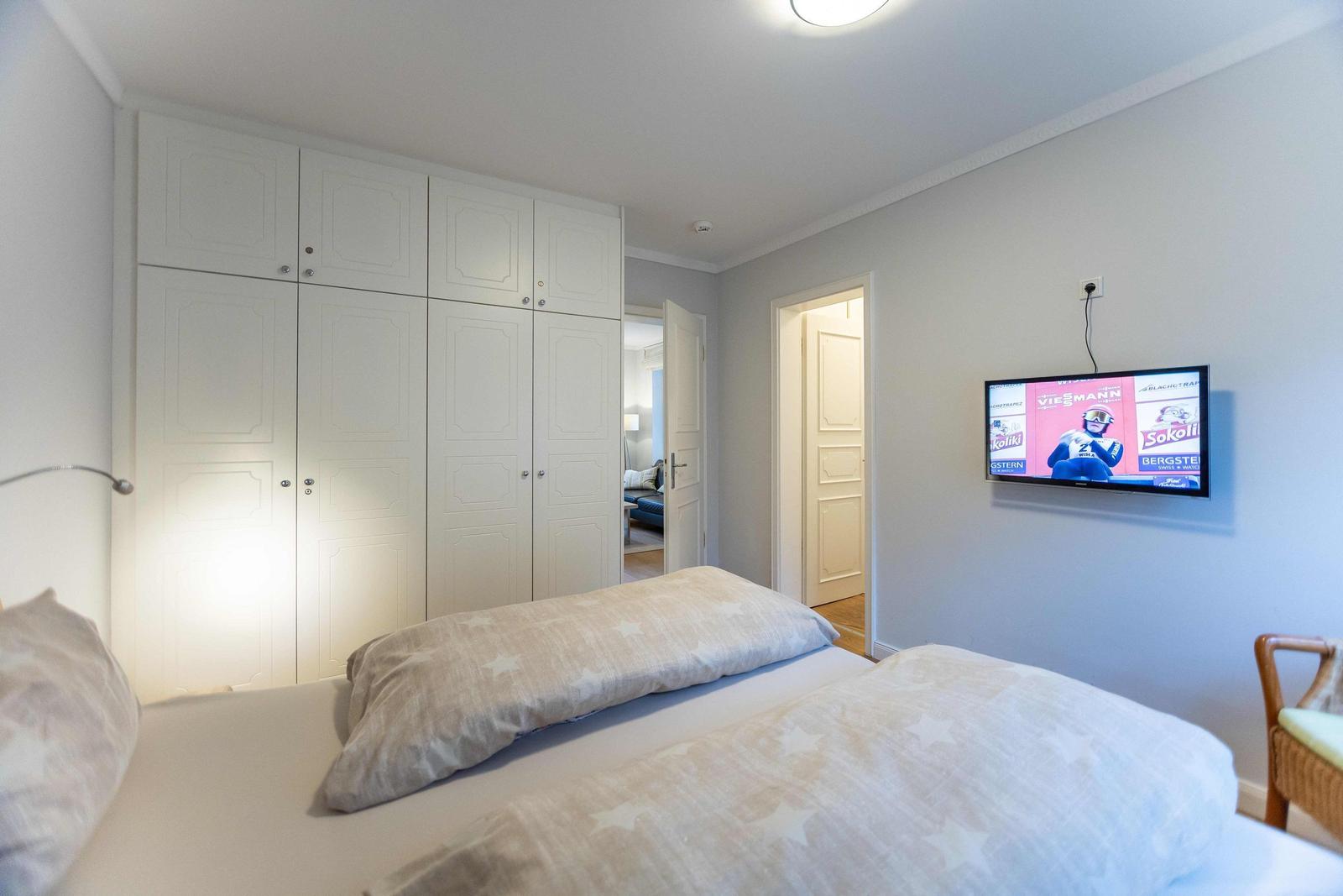 Schlafzimmer mit Doppelbett, TV und Blick in angrenzenden Raum mit Fenster.