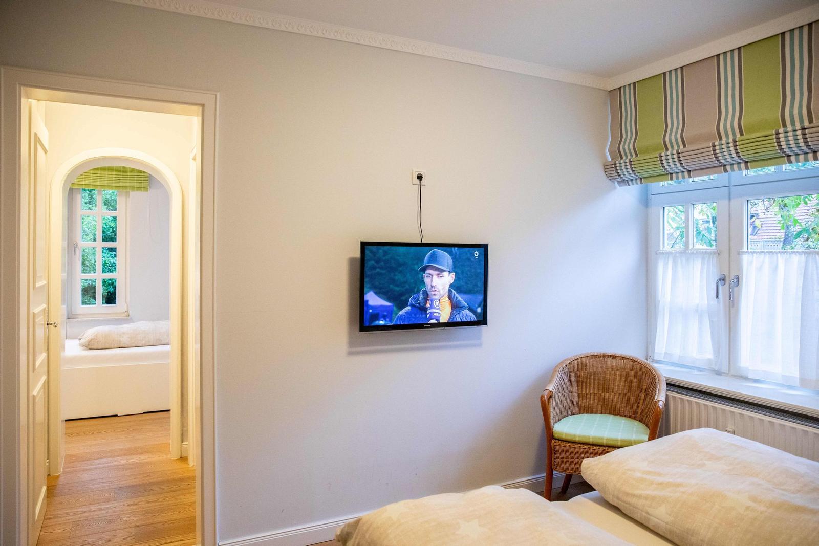 Zimmer mit Schreibtisch, Stuhl und zwei Fenstern mit grünen Rollos.