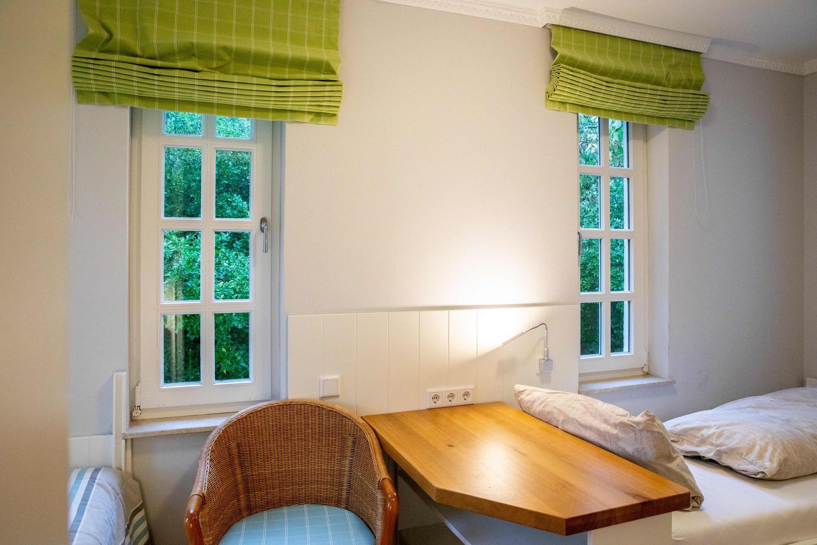 Ein Schlafzimmer mit einem Bett, einem Schreibtisch und einem Stuhl. Zwei Fenster mit Vorhängen.