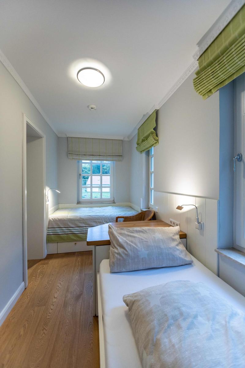 Ein Schlafzimmer mit Bett, Tisch und zwei Fenstern mit grünen Rollläden.